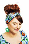 A woman from Izmir, smiling against a plain white background, wears the Cissi & Selma Hedda hårband humlesurr blå by BUTIK KUBIK. Her hairstyle is a large bun, and she complements her colorful floral top with matching vibrant earrings shaped like flowers. Her subtle makeup highlights her cheerful expression.