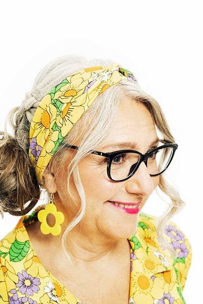 A woman with gray hair and glasses smiles, showcasing her vibrant BUTIK KUBIK Cissi & Selma Hedda hårband humlesurr gul made from ekologisk bomull with floral patterns that match her dress. Large yellow flower-shaped earrings complete her look against a simple white background.