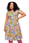 A woman with curly hair smiles in the Cissi och Selma Hilda dress Växtkraft Rosa from BUTIK KUBIK—a colorful, short-sleeve floral dress in bold orange, pink, purple, and white tones, paired with pink leggings.