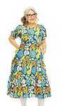 Smiling older woman with gray hair and glasses wearing the Cissi och Selma Ida klänning Blomklasar from BUTIK KUBIK, standing with hands in her pockets against a white background.