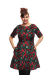 A woman with brown hair in an updo, wearing green earrings and the Cissi & Selma dress Irma Symfoni by BUTIK KUBIK, stands smiling against a white background with her hands in her dress pockets.