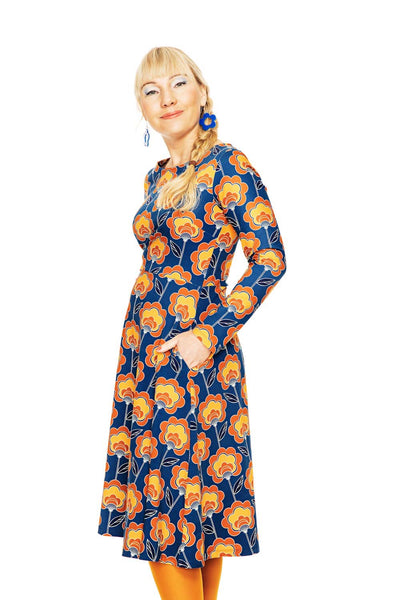 A blonde woman in pigtails wears the BUTIK KUBIK Cissi och Selma dress Kajsa Retroblom Blåbrun with orange tights and blue earrings, posing against a white background with her hands in her pockets.