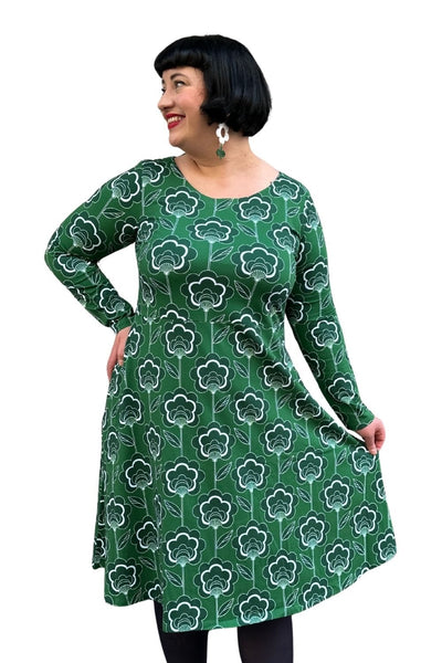 A woman with short dark hair wears the Cissi och Selma dress Kajsa Retroblom Grön from BUTIK KUBIK, featuring a green long-sleeve design with white floral print and side pockets. She smiles, looks sideways, holds her skirt, and wears white floral earrings.