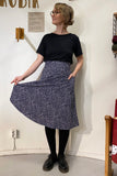 A person indoors wears a black T-shirt paired with the 50s-inspired Kubik skirt Twist Pearlsugar by BUTIK KUBIK. Holding the navy blue skirt with white dots in their left hand, they have glasses, short hair, black tights and shoes. Behind them is a sign and yarn on the wall.