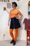 A person in a black cap, orange and white striped shirt, BUTIK KUBIK Kubik corduroy zip skirt nr 1, bright orange tights, and black shoes stands indoors by a wall and a chair draped with another similar striped shirt.