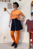 A person in a black cap, orange and white striped shirt, BUTIK KUBIK Kubik corduroy zip skirt nr 1, bright orange tights, and black shoes stands indoors by a wall and a chair draped with another similar striped shirt.