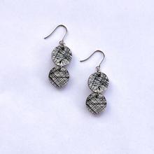 The Törmi Satu earrings by BUTIK KUBIK feature two round, black and white crisscross-etched discs in silver-tone surgical steel, shown against a plain white background.