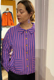 A woman stands indoors looking to the side, wearing the BUTIK KUBIK Kubik cotton shirt striped—an oversized, Sweden-inspired piece in purple and blue stripes with large pockets, crafted from Oeko-Tex cotton. Behind her, clothing hangs on the wall.