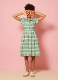 A woman in a green Palava Louise dress from BUTIK KUBIK stands against a pink background, holding two grapefruit halves over her eyes. She has curly hair pulled up and wears casual beige sneakers.
