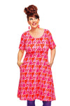 A smiling woman with red hair in an updo models the Cissi och Selma Magda dress Fridfull Rosa by BUTIK KUBIK, paired with pink earrings and purple leggings, standing against a white background with her hands in the dress pockets.