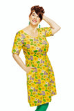 A smiling person wears the Cissi och Selma Magda dress Humlesurr gul by BUTIK KUBIK, adorned with green and purple flowers, green earrings, and green tights while posing confidently against a white background.
