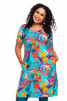A smiling woman with curly hair wears the Cissi och Selma Magda dress Kazoo Turkos by BUTIK KUBIK, featuring bold orange, purple, and red abstract florals on turquoise organic cotton. She stands with hands in pockets against a white background.