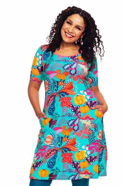 A smiling woman with curly hair wears the Cissi och Selma Magda dress Kazoo Turkos by BUTIK KUBIK, featuring bold orange, purple, and red abstract florals on turquoise organic cotton. She stands with hands in pockets against a white background.