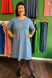 A smiling person with short dark hair stands indoors wearing the Cissi och Selma Magda dress Mört blå from BUTIK KUBIK, black tights, and black shoes. Colorful dresses hang on racks with a green wall in the background.