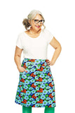 Smiling woman with gray hair and glasses wears a white top, green tights, and the BUTIK KUBIK Cissi och Selma Majken skirt Humlesurr Blå in organic cotton jersey, standing with hands in her skirt pockets against a white background.