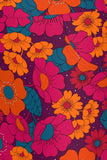 The Cissi och Selma Majken skirt Klang by BUTIK KUBIK features a vibrant floral pattern with large orange and pink flowers, teal centers, and green leaves on a dark purple background. Crafted from ekologisk bomullstrikå, it offers a lively design and fits normal i storleken.