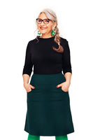 Smiling woman with long gray braided hair, glasses, green earrings, and black top wears the Cissi och Selma Majken skirt Mörkgrön from BUTIK KUBIK, hands in pockets, standing against a plain white background.