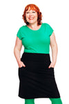 Woman with red, curly hair smiling, wearing a bright green top and the Cissi och Selma Majken skirt svart by BUTIK KUBIK, with green tights and hands in pockets, standing against a white background.