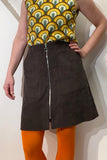 A person stands with one hand on their hip, wearing a retro rainbow top, a BUTIK KUBIK Kubik brown cotton corduroy zip skirt, and bright orange tights against a plain white wall. All materials are Oeko-Tex certified for sustainability.