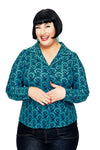 A smiling woman with short black hair, wearing the Cissi och Selma blus Matilda Miniblom Blå from BUTIK KUBIK and dark jeans, stands against a white background with her hands gently clasped together.