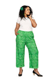 A woman with curly hair wears a white t-shirt, Cissi och Selma Molly pants Ängsblom grön by BUTIK KUBIK, black heels, and a green floral hair accessory. She smiles with one hand on her face and the other in her pocket, against a white background.