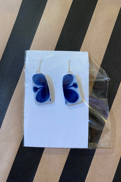 A pair of Saved & remade earrings by BUTIK KUBIK, featuring blue and white rectangles made from återbrukat porslin with silverpläterade örhängen hooks, shown on a white card partially wrapped in clear plastic on a striped tan and black surface.
