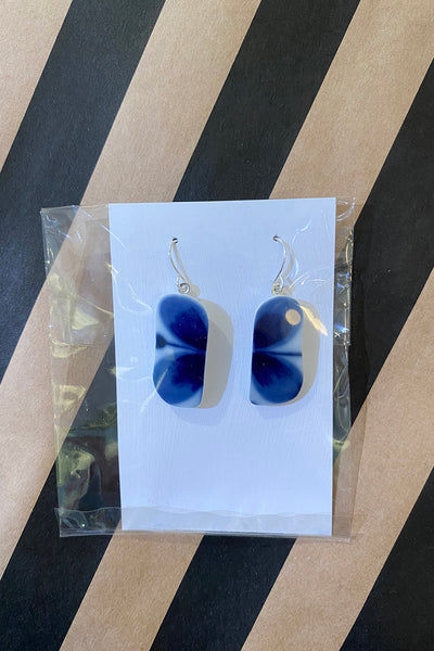 A pair of Saved & remade earrings by BUTIK KUBIK, featuring rectangular shapes in blue and white floral designs from återbrukat porslin, displayed on a white card in a clear bag against a diagonal black and tan striped background.