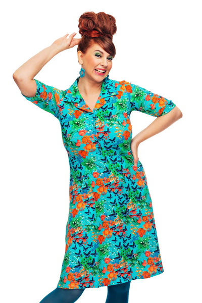 Smiling woman with bright red updo, teal earrings, blue tights, and the Cissi och Selma dress Monica Fjärilar by BUTIK KUBIK— a vibrant 70s-inspired shirt dress featuring orange, green, and blue butterfly patterns on white.
