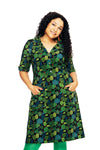 Woman with long curly hair in a 70s-inspired "Monica Listig" dress by BUTIK KUBIK, made of organic cotton jersey, green leaf pattern, buttons, short sleeves, hands in pockets, smiling against a white background.