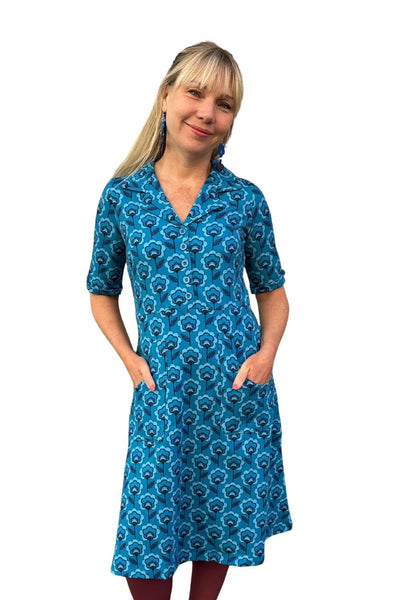 En leende kvinna med långt blont hår bär en blå Cissi och Selma dress Monica Miniblom från BUTIK KUBIK, en 70-talsinspirerad skjortklänning i ekologisk bomull med korta ärmar och fickor, mot vit bakgrund.