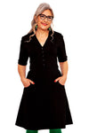 A smiling woman with gray hair, glasses, and green earrings wears the "Monica Jeans Svart" dress by BUTIK KUBIK—knee-length, black, with buttons and short sleeves. She stands with hands in her pockets against a white background.