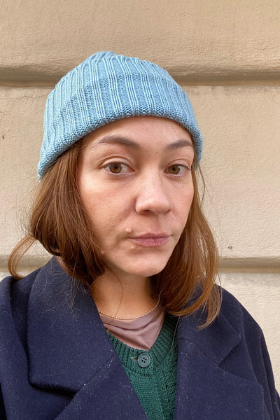 A woman with light brown hair wears a Mariedal wool beanie aqua from BUTIK KUBIK, a dark coat, and a green sweater. She stands in front of a beige wall, looking directly at the camera with a neutral expression.