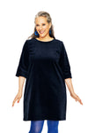 A smiling woman with gray and dark hair wears the BUTIK KUBIK Cissi & Selma dress Olivia Corduroy Blå, styled with blue tights and earrings, standing against a white background.