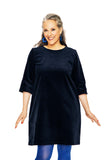 A smiling woman with gray and dark hair wears the BUTIK KUBIK Cissi & Selma dress Olivia Corduroy Blå, styled with blue tights and earrings, standing against a white background.