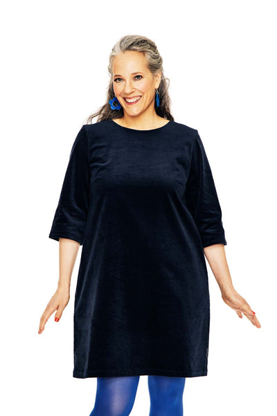 A smiling woman with gray and dark hair wears the BUTIK KUBIK Cissi & Selma dress Olivia Corduroy Blå, styled with blue tights and earrings, standing against a white background.