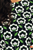 A close-up of a person wearing the Cissi & Selma dress Olivia Retroblom svart from BUTIK KUBIK, featuring a bold repeating pattern of white flowers with green stems. The person's wavy dark hair is partially visible on the left.