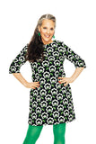 A smiling woman with long, dark hair and gray streaks poses in the Cissi & Selma dress Olivia Retroblom svart from BUTIK KUBIK, featuring a white flower pattern, bright green tights, earrings, and ring against a white background.