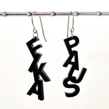 Two black Favoretro Earrings Fikapaus Plain Black by BUTIK KUBIK, crafted from laserskuren akryl in stacked letter shapes, hang from a metal rod. One earring spells “FIKA” vertically, the other “PAUS.” Both svarta örhängen feature silver hooks in kirurgiskt stål and bold, playful lettering.
