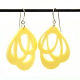 The Favoretro Earrings Loops lemon haze by BUTIK KUBIK are yellow, teardrop-shaped acrylic earrings with abstract cutouts, hanging from a silver surgical steel bar against a white background.