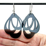 A close-up of a hand holding Favoretro Earrings Loops Smokey black by BUTIK KUBIK, modern acrylic hoops with abstract cut-outs, hanging from a black rod against a white background.