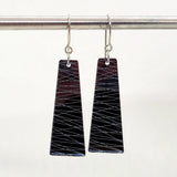 A pair of Favoretro earrings sketch by BUTIK KUBIK, featuring trapezoid-shaped thundercloud gray designs crafted from laser-cut plexiglass with silver zigzag lines, hang from a surgical steel rod against a plain white background.