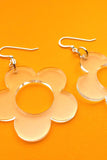 The Cissi & Selma Örhängen Solo Vatten by BUTIK KUBIK are clear, acrylic, flower-shaped earrings with silver metal hooks. Their retro design features five rounded petals and a hollow center, ideal for unique style lovers, shown on a bright orange background.