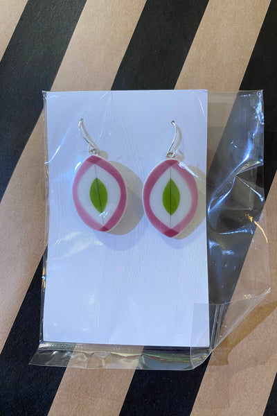 Saved & remade earrings by BUTIK KUBIK: onion-shaped with pink and white rings, green center, crafted from återbrukat porslin. Displayed on a white card, partially wrapped in clear plastic on a striped background.