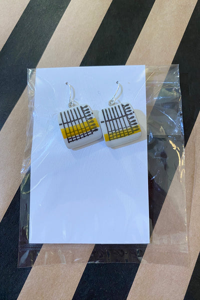 BUTIK KUBIK's Saved & Remade earrings are square with a white base, black lines, and yellow-gray rectangular patterns made from återbrukat porslin, displayed on a white card in a clear plastic sleeve on a diagonal striped surface.