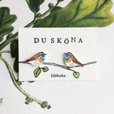 A pair of Du sköna earrings Blåhake by BUTIK KUBIK, featuring hand-painted blue and brown hues inspired by the blåhake, are displayed on a DU SKÖNA card with illustrated green leaves and branches.