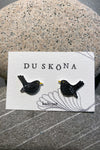 A pair of black Du sköna bird earrings by BUTIK KUBIK, featuring yellow beaks, are displayed on a card labeled DU SKÖNA and koltrast, resting against smooth, speckled gray stones.