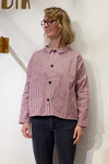 A woman with wavy blonde hair and glasses wears the BUTIK KUBIK Kubik Overshirt Strömma Kadett blåröd, featuring red, white, and blue vertical stripes, over dark jeans while standing against a plain white wall.