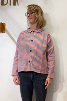 A woman with wavy blonde hair and glasses wears the BUTIK KUBIK Kubik Overshirt Strömma Kadett blåröd, featuring red, white, and blue vertical stripes, over dark jeans while standing against a plain white wall.
