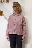 A woman with wavy blonde hair and glasses wears the BUTIK KUBIK Kubik Overshirt Strömma Kadett blåröd, featuring red, white, and blue vertical stripes, over dark jeans while standing against a plain white wall.