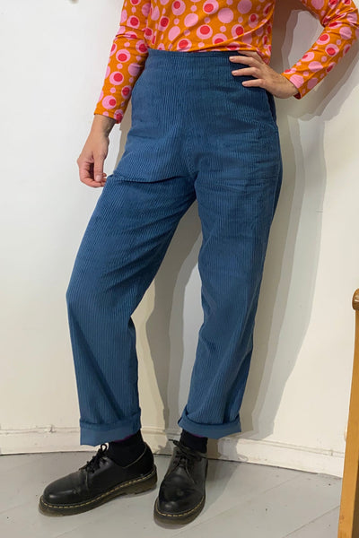 A person stands against a white wall wearing BUTIK KUBIK's high-waisted Kubik corduroy trousers, black shoes, and a long-sleeve orange and pink circle-patterned top. Only the lower half of their face is shown.